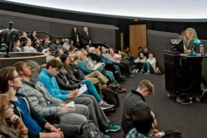 People sitting in an auditorium with a woman speaking from a podium at the front of the room