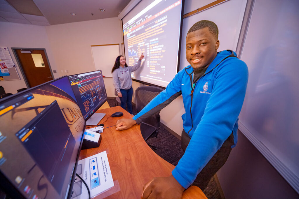 One student at a desktop computer and another student in the background by a large screen with information on the financial markets